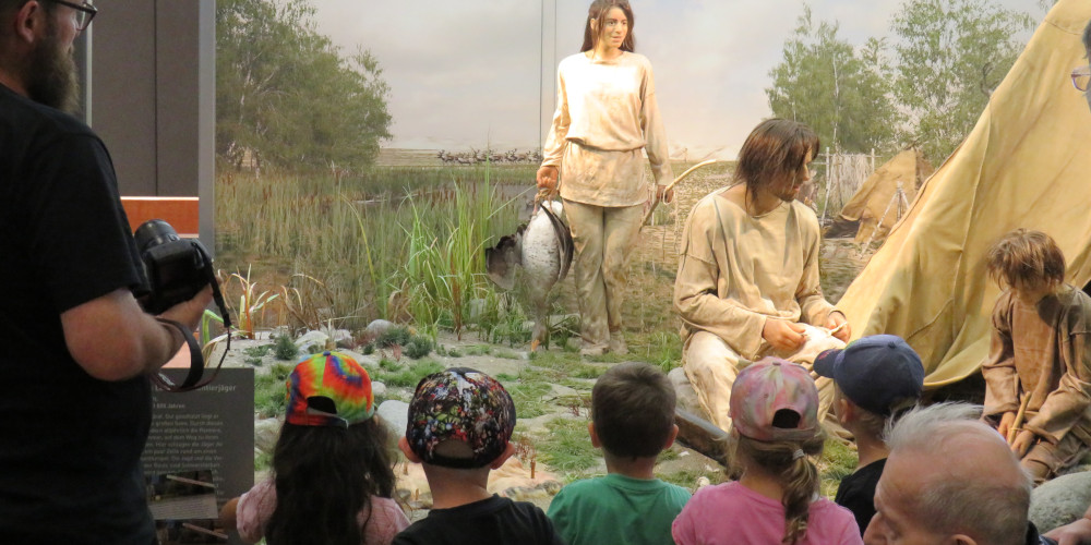 Kinder betrachten eine Urzeitszene mit Menschen im Museum, am linken Rand eine erwachsene Begleitperson.