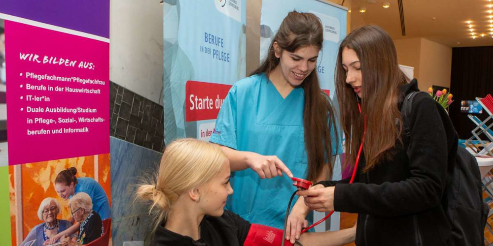 Auszubildende in Berufskleidung mit zwei Mädchen bei Blutdruckmessen vor Werbebannern
